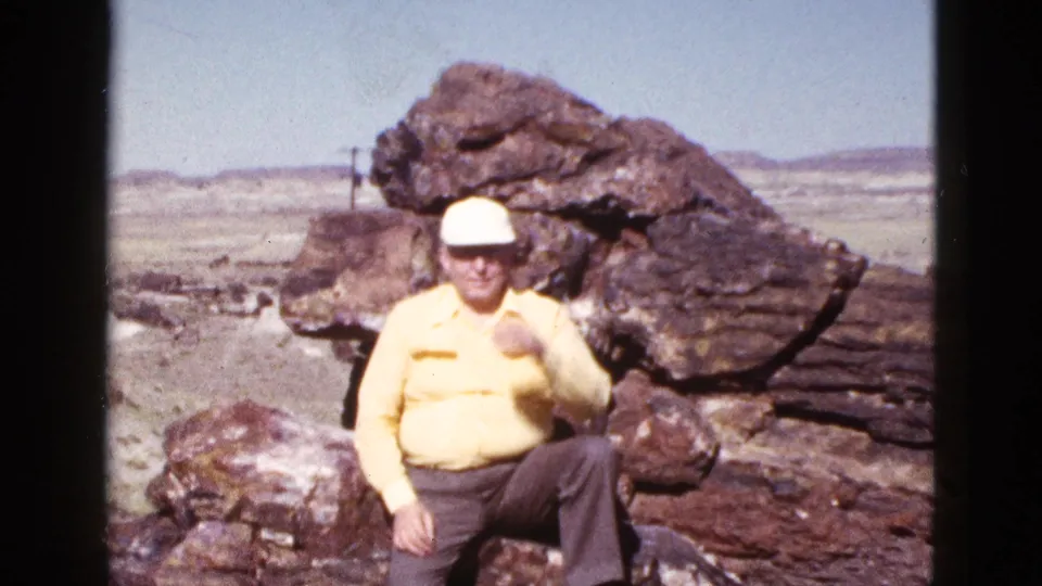 Older man in a yellow shirt and white cap sits against large rocks in the Arizona desert, facing the camera.
