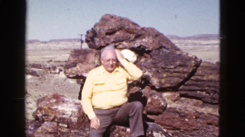 In the Arizona desert, the seated man in a yellow shirt raises his white cap while posing among large rocks.