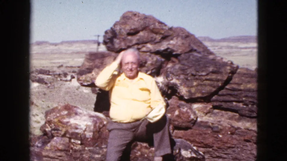 Seated older man in a yellow shirt strokes his hair beside large rocks in the 1953 Arizona desert footage.