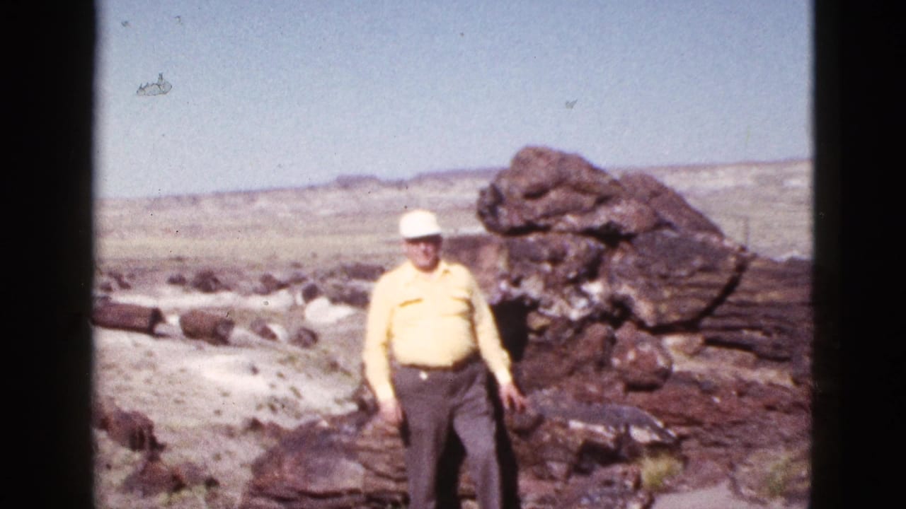 In 1953 Arizona home-movie footage, an elderly man in a yellow shirt and white cap stands near dark rock formations in a desert landscape.