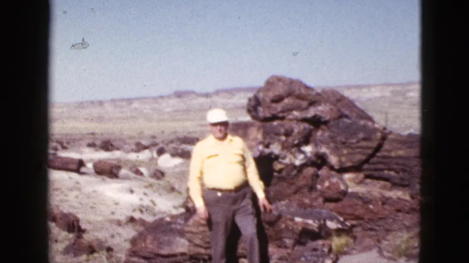 In 1953 Arizona home-movie footage, an elderly man in a yellow shirt and white cap stands near dark rock formations in a desert landscape.
