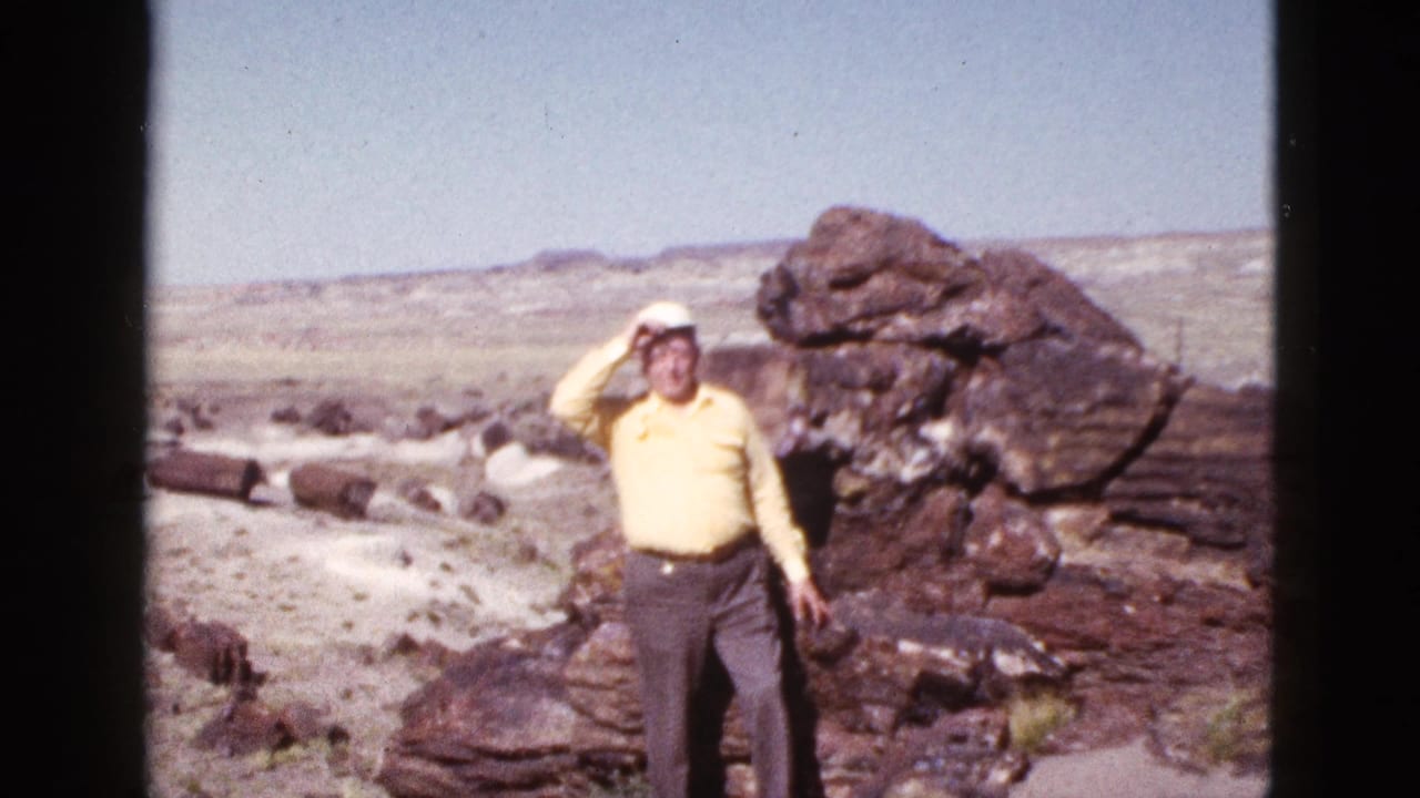 Elderly man touches his white cap while standing beside desert rock formations.