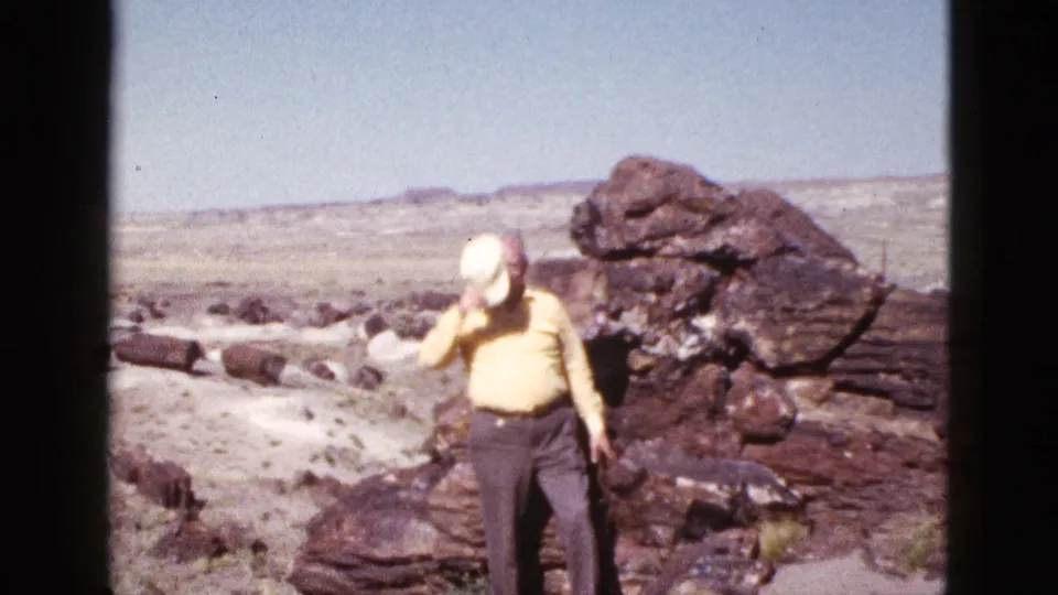 The elderly man lowers his gaze while handling his white cap, framed by dark desert rocks and open Arizona sky.