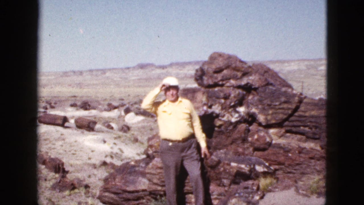 1953 Arizona home-movie frame showing an adult man in a yellow shirt posing near large desert rocks and shielding his eyes from sun.