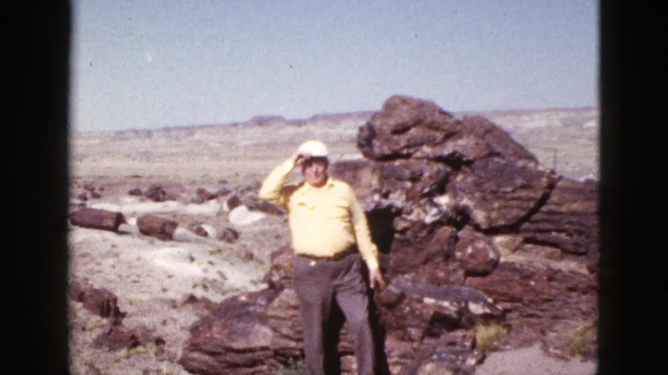 1953 Arizona home-movie frame showing an adult man in a yellow shirt posing near large desert rocks and shielding his eyes from sun.