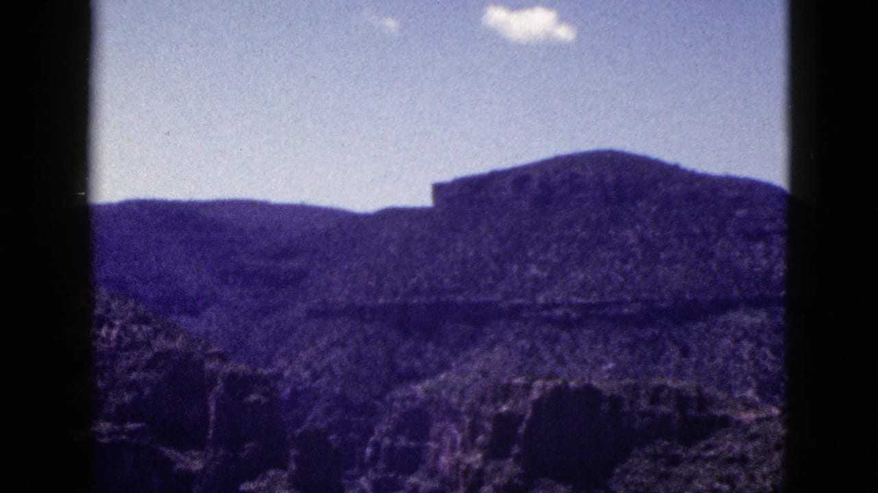 1953 New Mexico 8mm frame showing purple-toned desert mountains, rocky foreground, pale sky, and a small white cloud within dark film borders.