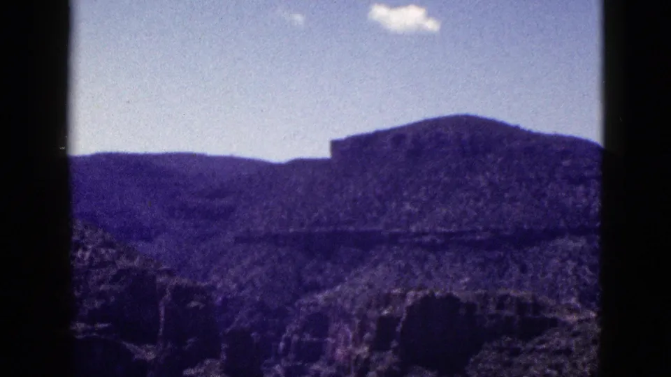 1953 New Mexico 8mm frame showing purple-toned desert mountains, rocky foreground, pale sky, and a small white cloud within dark film borders.