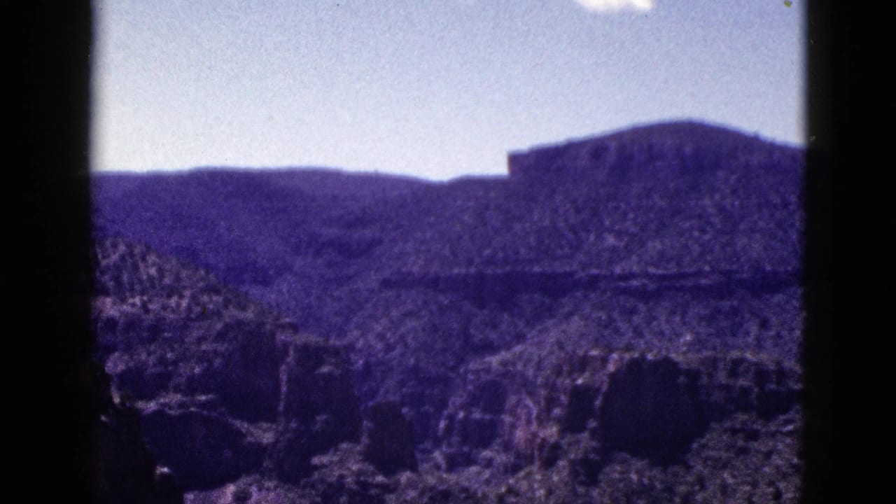 1953 New Mexico home-movie frame with a layered ridge, rough desert rock, pale sky, and strong purple color shift from the 8mm scan.