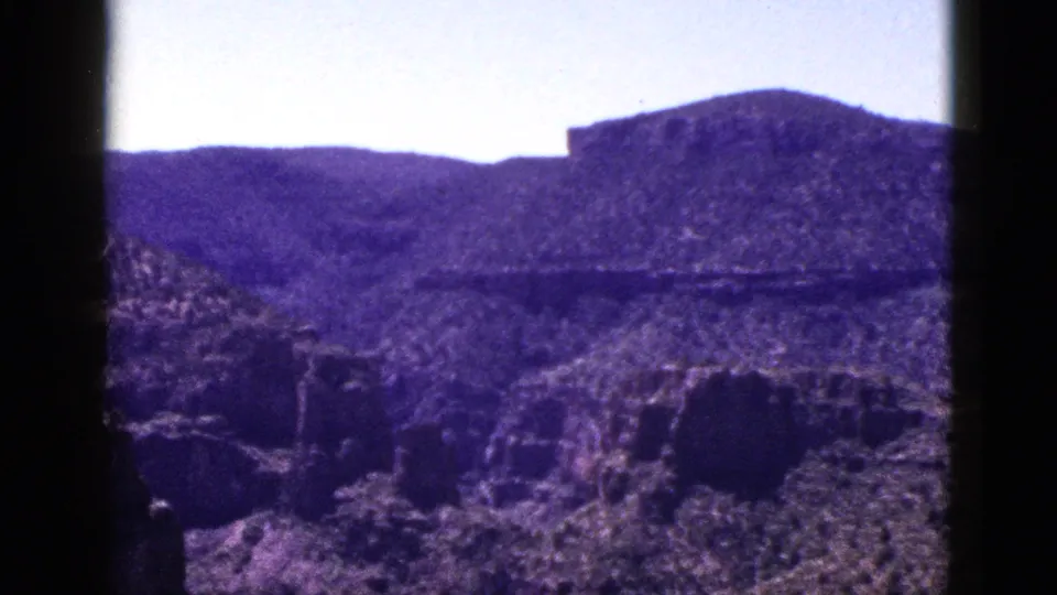 1953 New Mexico 8mm frame showing a closer desert hillside, rough rock formations, pale sky, and visible home-movie grain.
