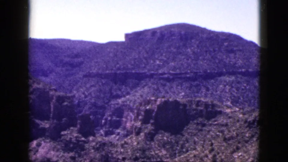 1953 New Mexico home-movie frame with mesa-like hills, rough foreground rock, pale sky, and dark 8mm projection borders.