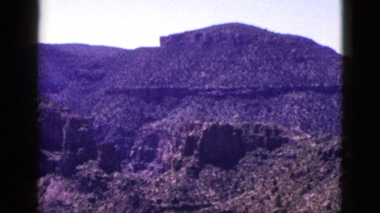 1953 New Mexico 8mm frame showing rugged canyon walls, arid slopes, distant mesa landforms, and bright sky with dark film edges.