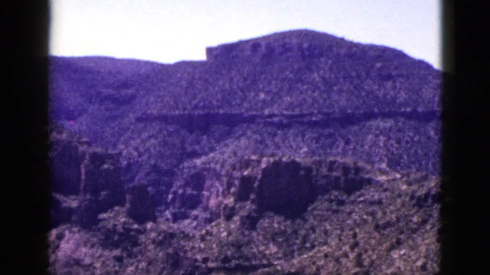 1953 New Mexico 8mm frame showing rugged canyon walls, arid slopes, distant mesa landforms, and bright sky with dark film edges.