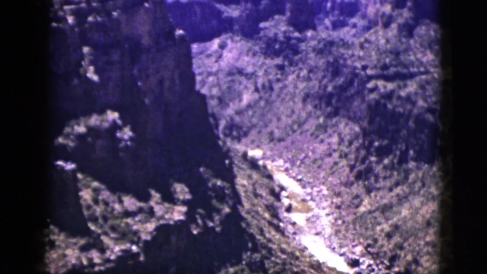 1953 New Mexico 8mm frame with a steep rock wall, high-desert vegetation, and a pale snowmelt stream crossing the valley.
