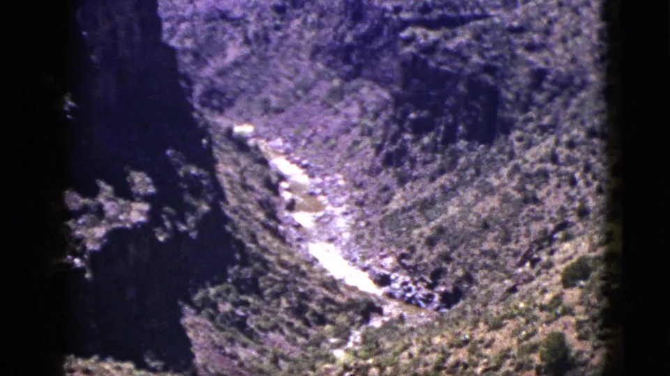 1953 New Mexico home-movie frame of a canyon river seen from above, bordered by rugged cliffs and dense vegetation.