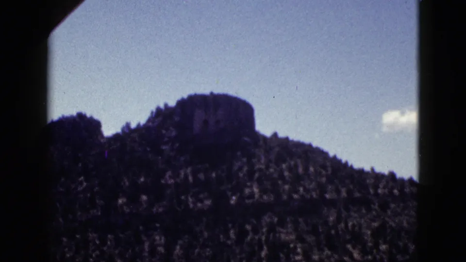 1953 New Mexico 8mm frame showing a distant mesa or butte above forested terrain beneath a clear blue sky.