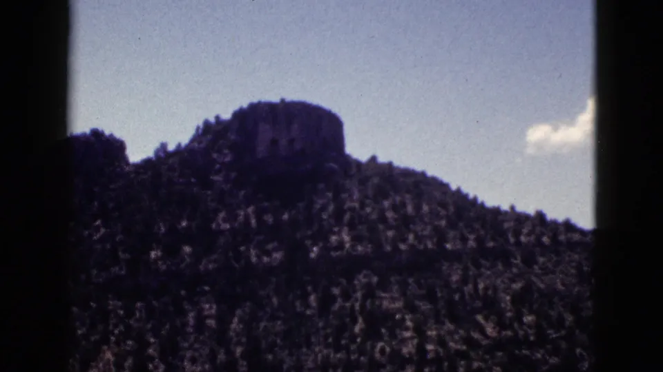 1953 New Mexico home-movie frame of a mesa or butte above a forested ridge, framed by blue sky and small cloud.