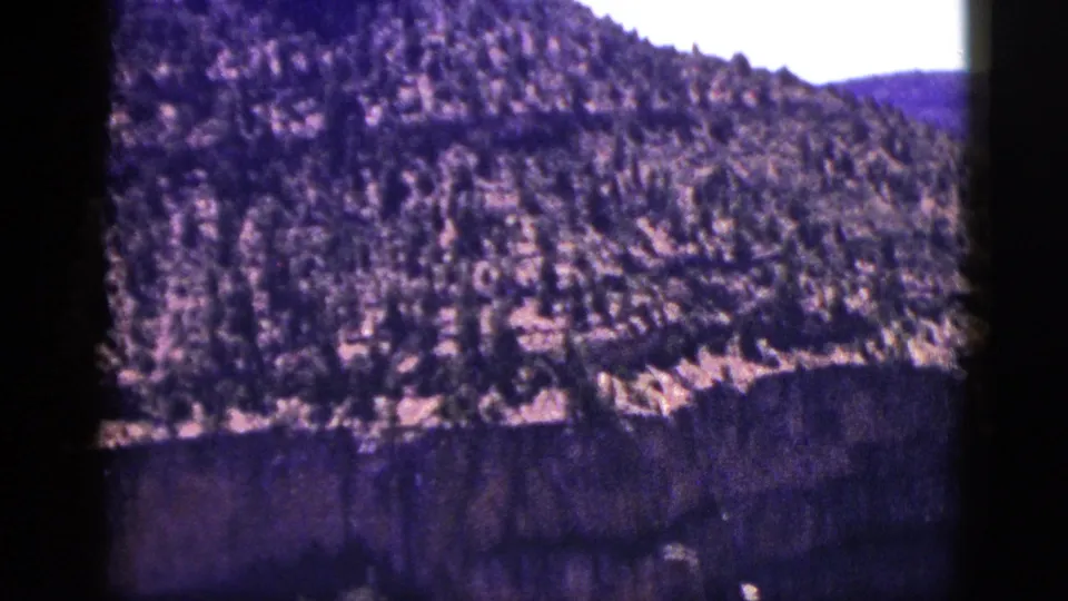 1953 New Mexico home-movie frame with a scrub-covered slope descending toward a dark layered cliff face.