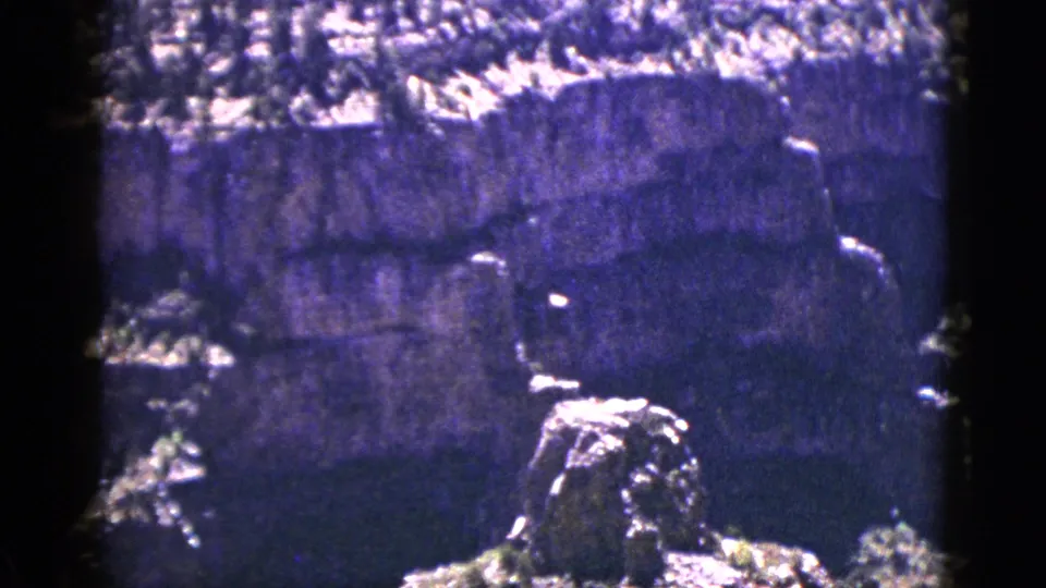 1953 New Mexico frame showing a shadowed cliff wall and bright foreground rocks within rugged mountain terrain.