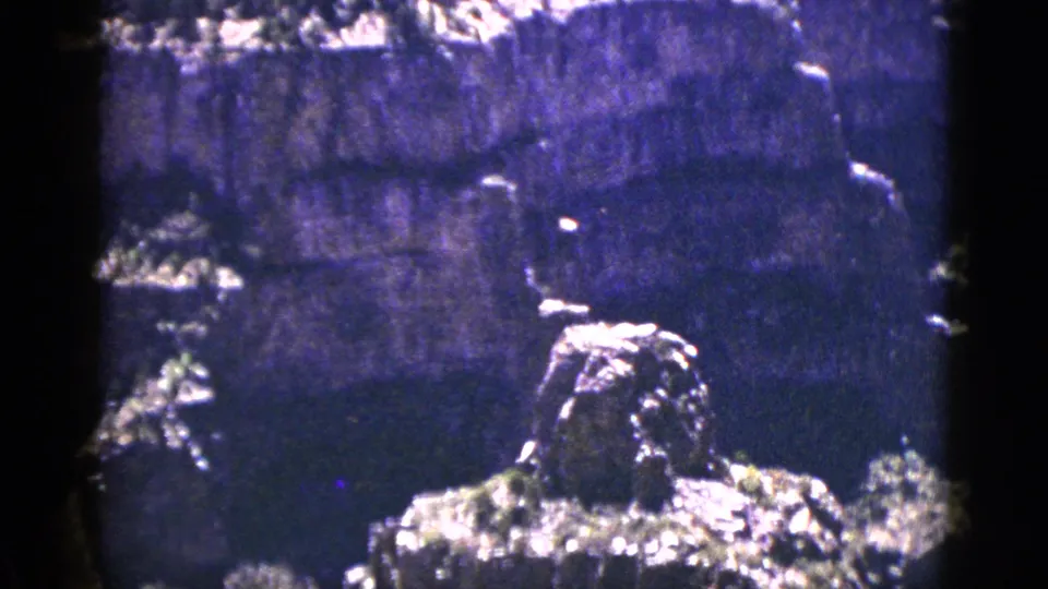 1953 8mm frame of a rugged canyon rock formation in New Mexico, with shrubs and heavy film grain visible.