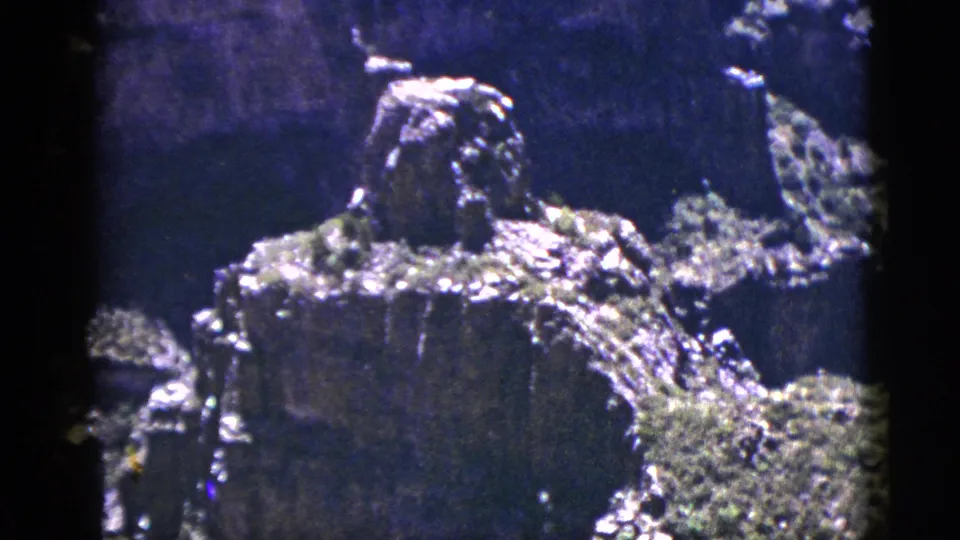 Close 1953 8mm canyon frame showing textured rock, a central outcrop, shrubs, and strong blue-purple color shift.