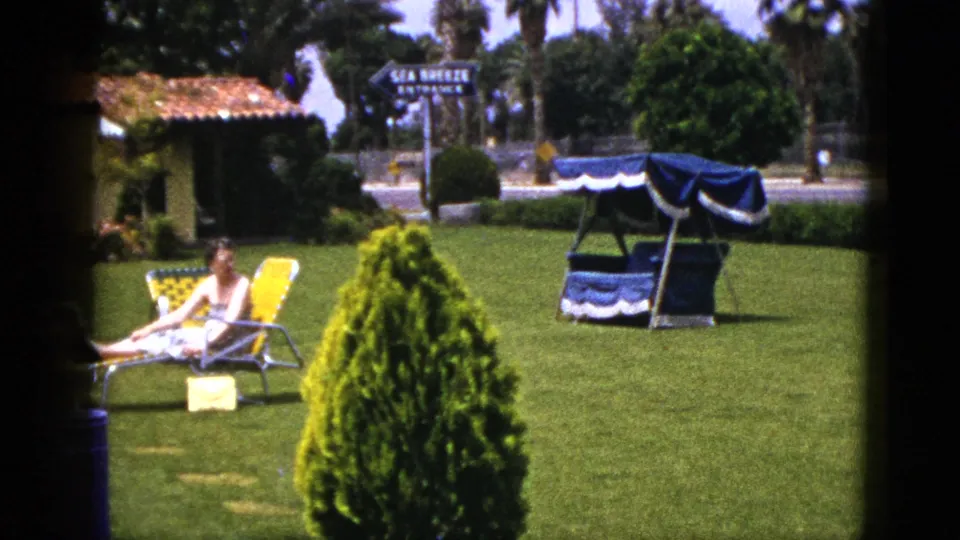 1953 resort lawn scene with a woman reclining near a yellow lounge chair, blue canopy swing, palm trees, and a Sea Breeze entrance sign.