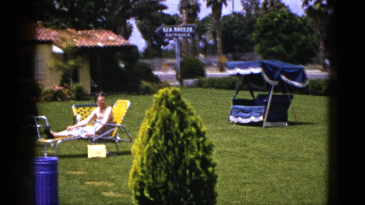 1953 New Mexico 8mm frame of a woman reclining on a yellow lawn chair in a maintained yard with a blue can, shrub, canopy swing, and entrance sign.