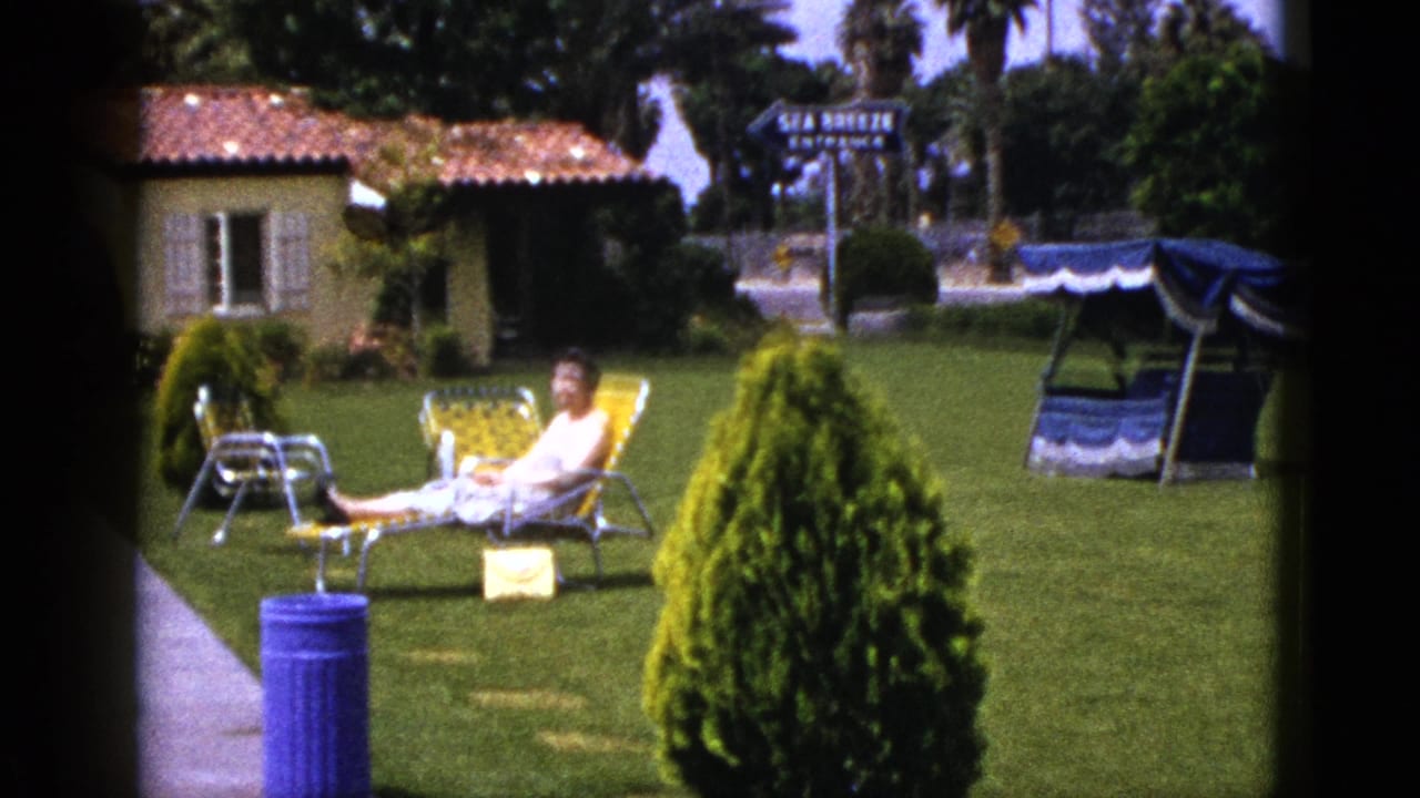 Woman seated in a yellow lounge chair on grass, with a blue can, shrub, small building, and canopy swing behind her.