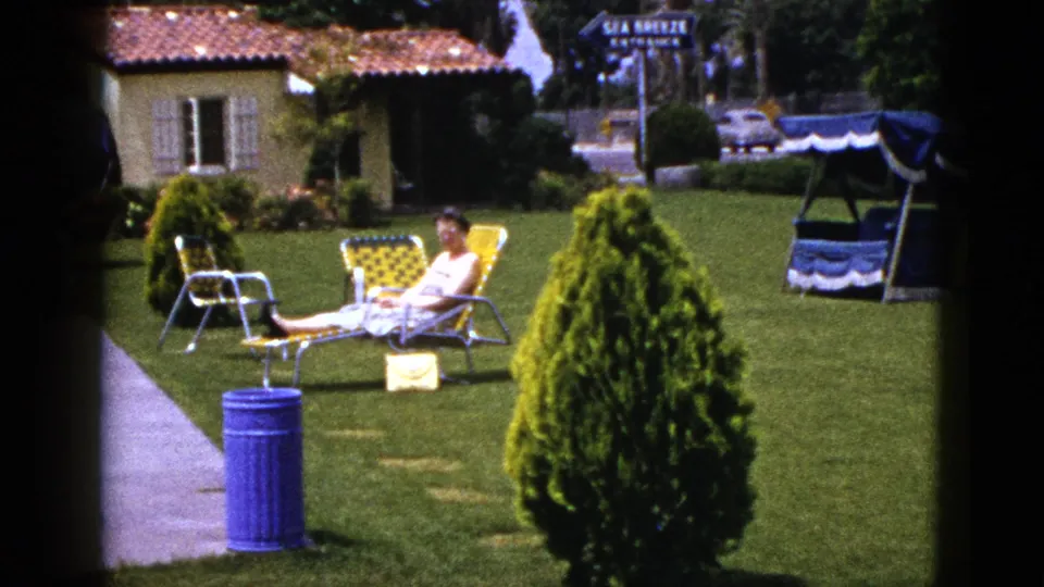 1953 New Mexico 8mm still of a woman on a yellow lawn chair, seen across grass with a conical shrub, blue can, bungalow, and roadside sign.