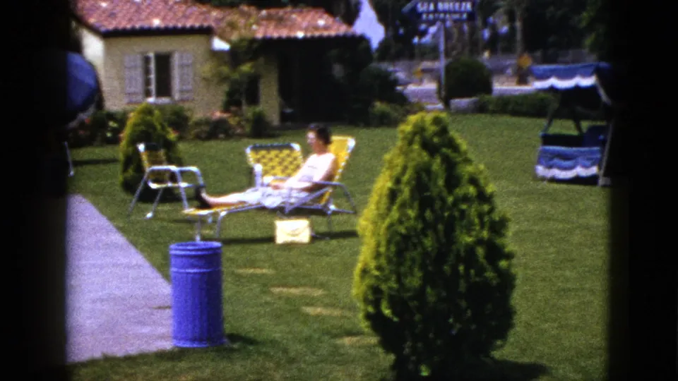 1953 New Mexico home-movie frame of a woman reclining in a yellow chair near a walkway, with a manicured lawn, shrub, blue can, and canopy swing.