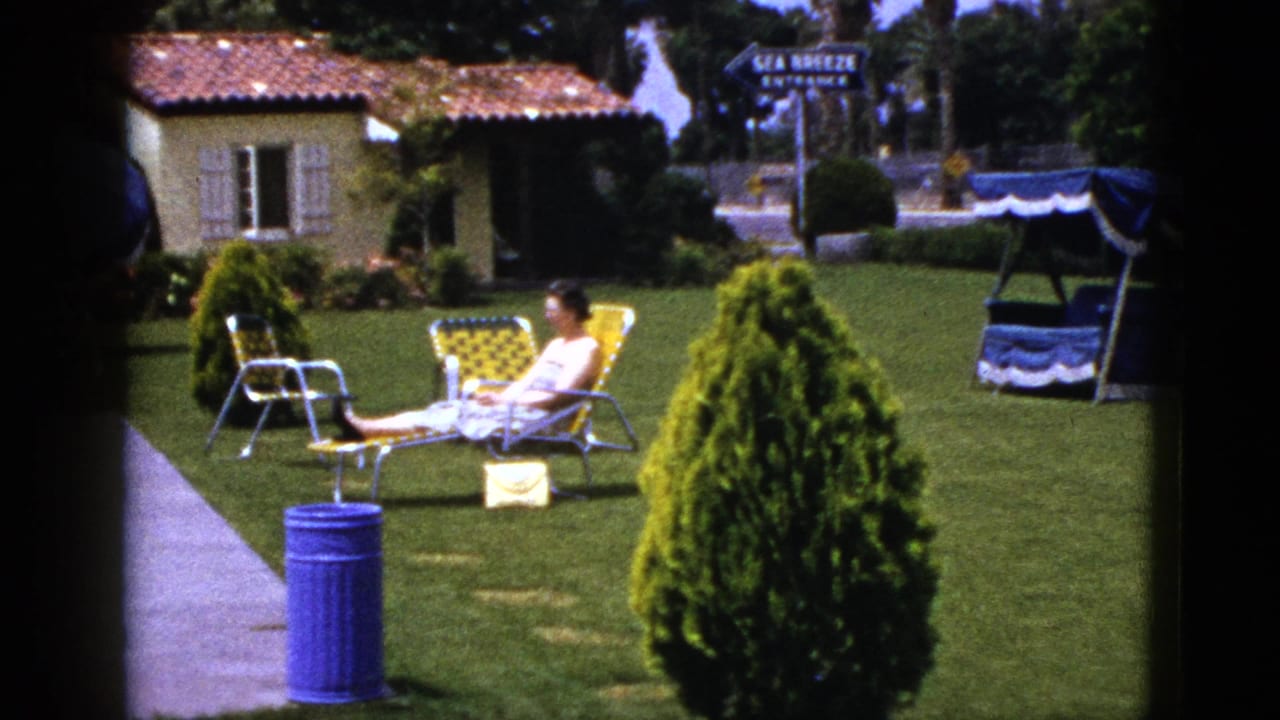 Woman reclines on a yellow lounge chair on a green lawn near a blue trash can and stucco building.