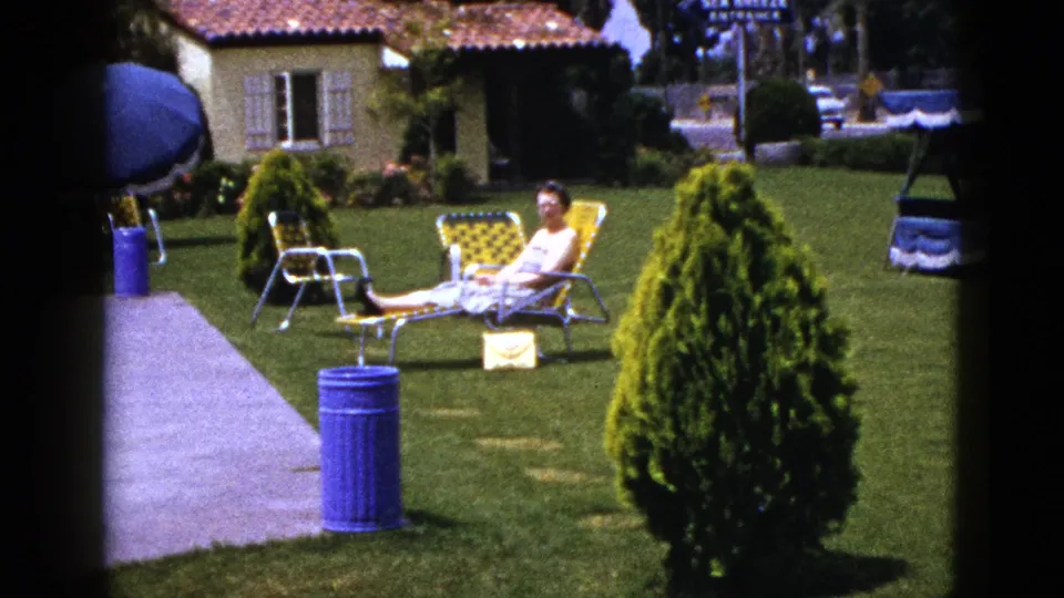 1953 home-movie frame showing a woman seated in a garden lounge chair near a walkway, clipped shrubs, blue trash can, and tiled-roof stucco structure.