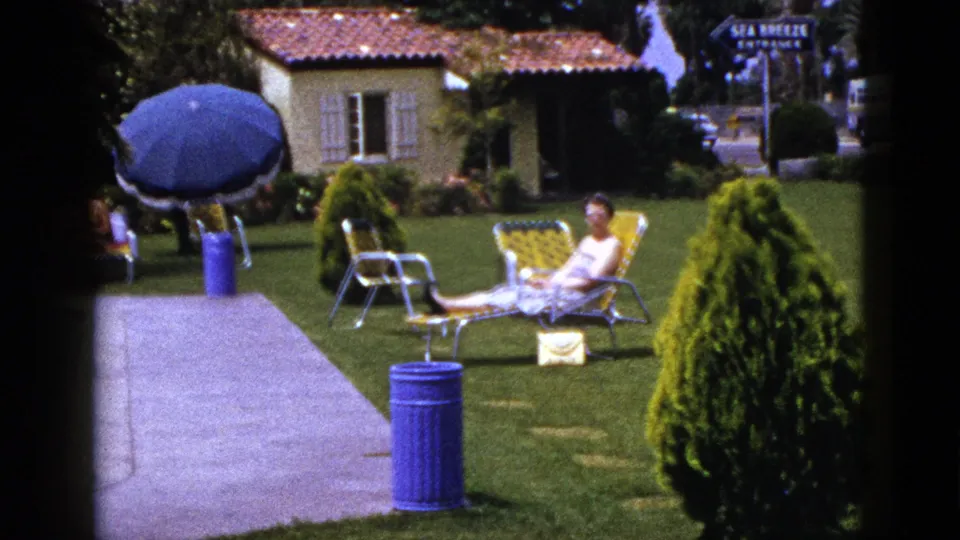 1953 8mm frame with a woman reclining in a landscaped garden as a blue umbrella, paved walk, shrubs, and stucco building fill the scene.