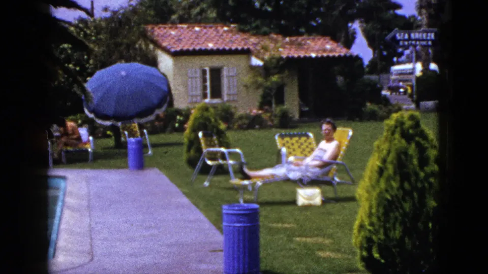 1953 home-movie frame of a woman lounging on a garden chair near a blue umbrella and walkway, with a stucco building and sign in the background.