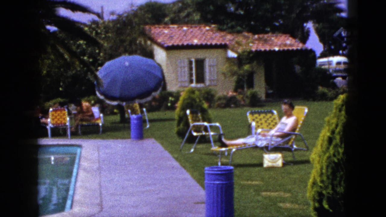Woman lounging beside a small pool while others sit on lawn chairs near a blue umbrella.