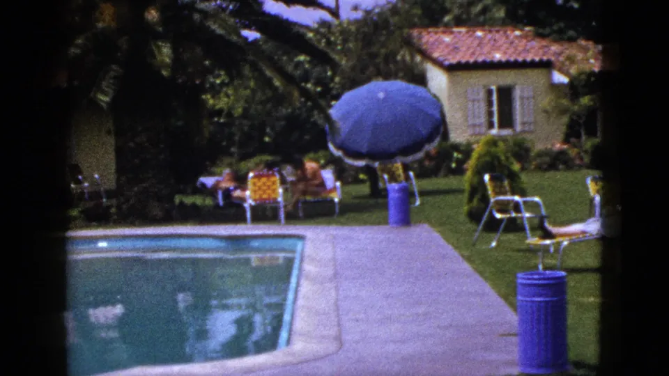 1953 New Mexico 8mm frame with the pool more prominent in foreground, adults relaxing on lawn chairs, and a tiled-roof house behind trees.