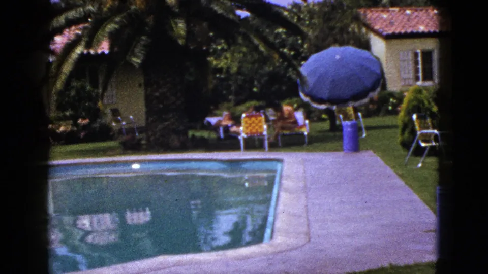 1953 New Mexico home-movie frame showing a small pool, clipped lawn, blue umbrella, yellow chairs, and adults relaxing near a modest home.