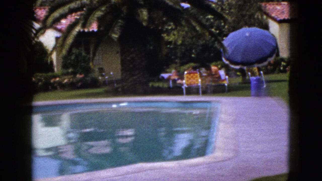 1953 8mm home-movie view in New Mexico of a poolside patio, stucco house, garden, blue umbrella, and distant people seated outdoors.