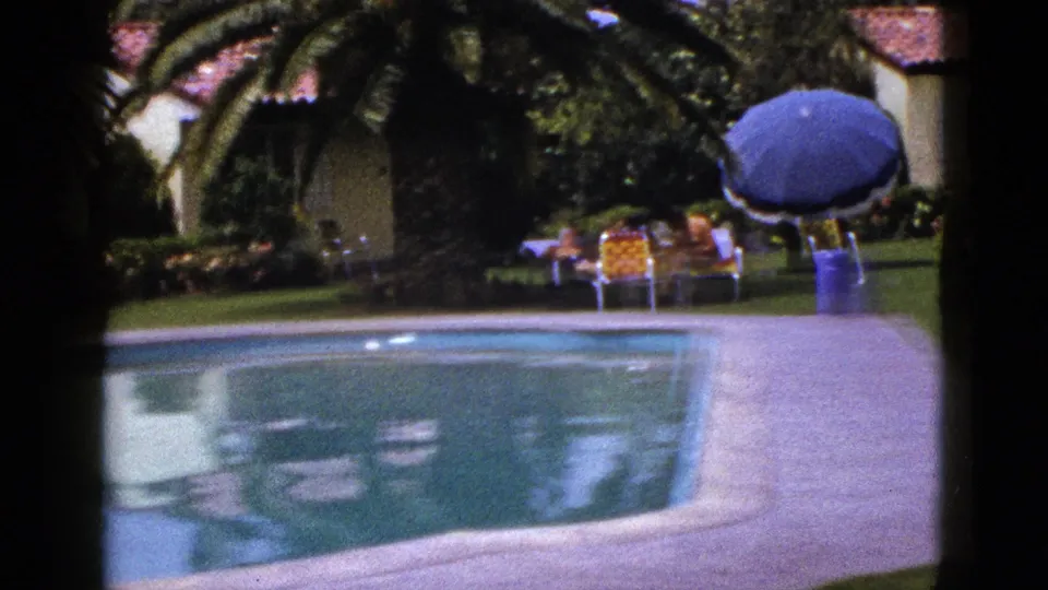 1953 8mm home-movie view in New Mexico of a poolside patio, stucco house, garden, blue umbrella, and distant people seated outdoors.