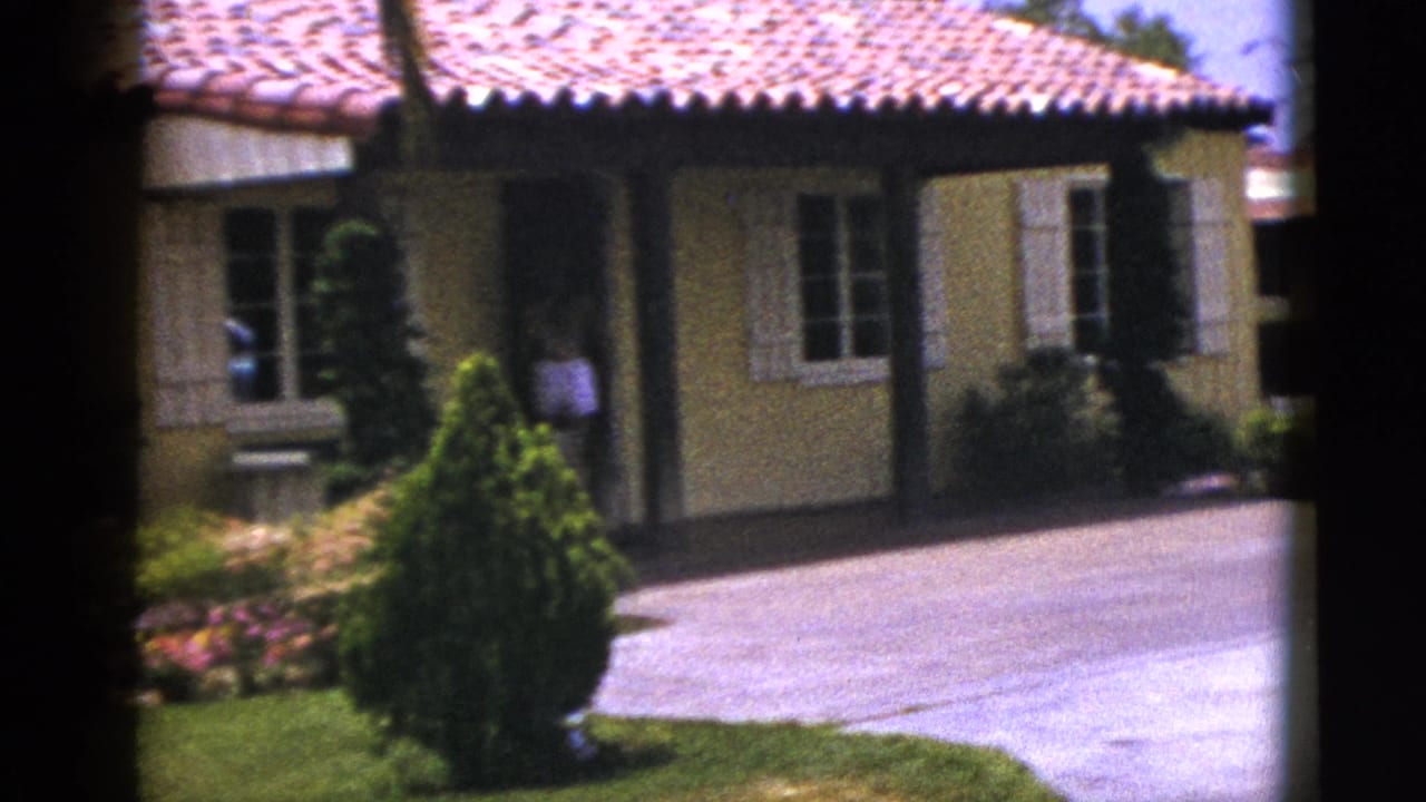 Stucco house exterior with red tile roof, shuttered windows, driveway, shrubs, and lawn.