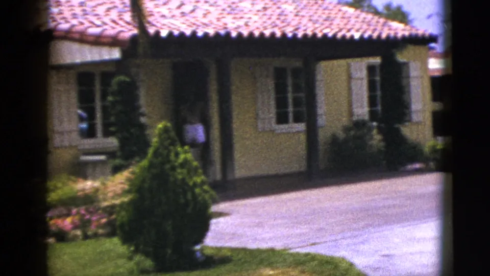 1953 New Mexico home-movie frame showing a stucco residence with red tile roof, white shutters, driveway, shrubs, and bright lawn.