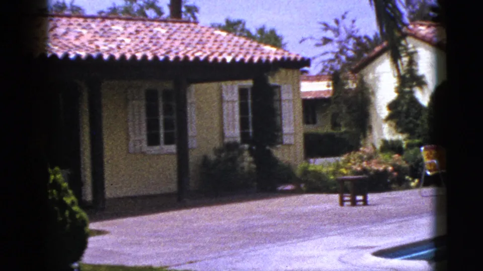 1953 8mm view of a New Mexico stucco house, pool deck, garden plantings, patio furniture, and shuttered windows in strong sunlight.