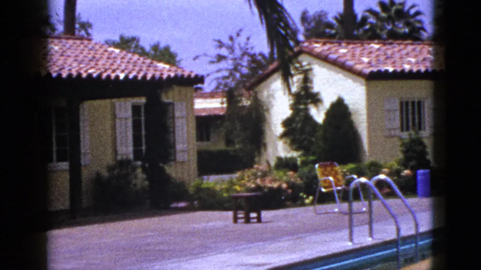 1953 New Mexico 8mm frame with pool ladder in the foreground, stucco house and garden beyond, plus patio seating and palm foliage.