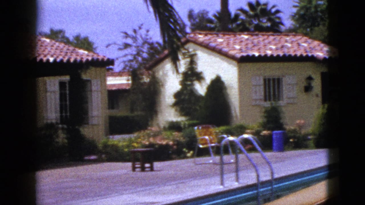 1953 New Mexico 8mm frame showing a backyard pool, concrete deck, stucco house with red tile roof, palms, chairs, and shrubs.