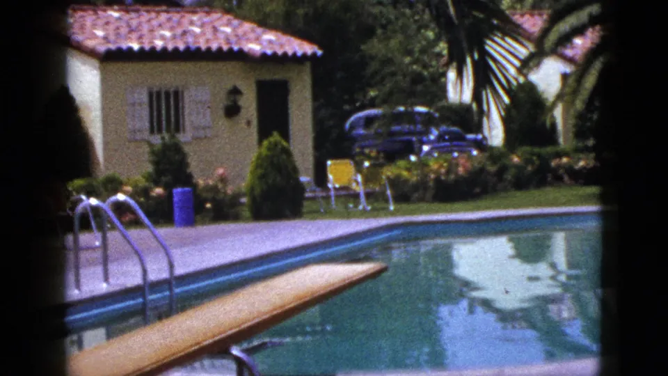1953 New Mexico home-movie frame of a backyard pool with diving board, metal ladder, stucco house, palms, shrubs, chairs, and blue bin.