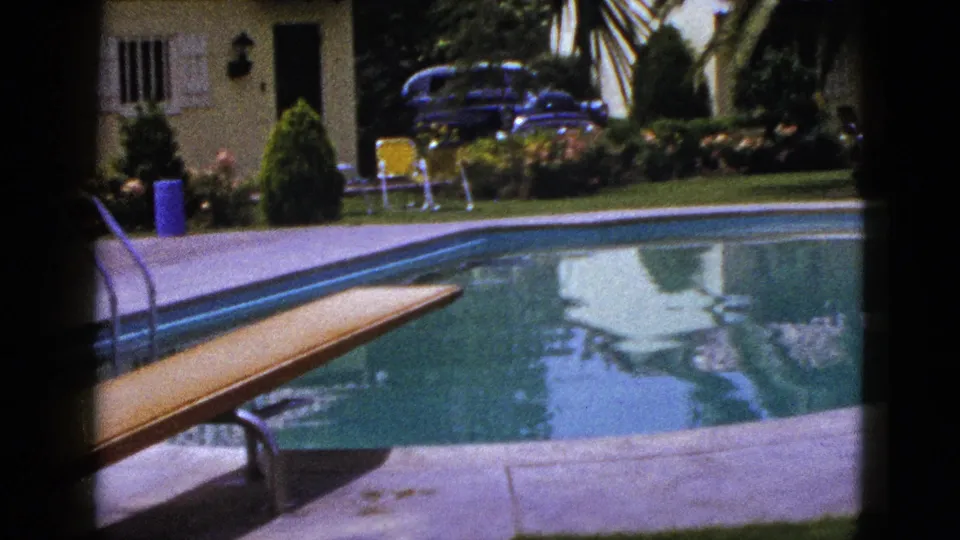 1953 New Mexico backyard pool scene with diving board over water, stucco house, palms, yellow chair, shrubs, blue bin, and parked car.