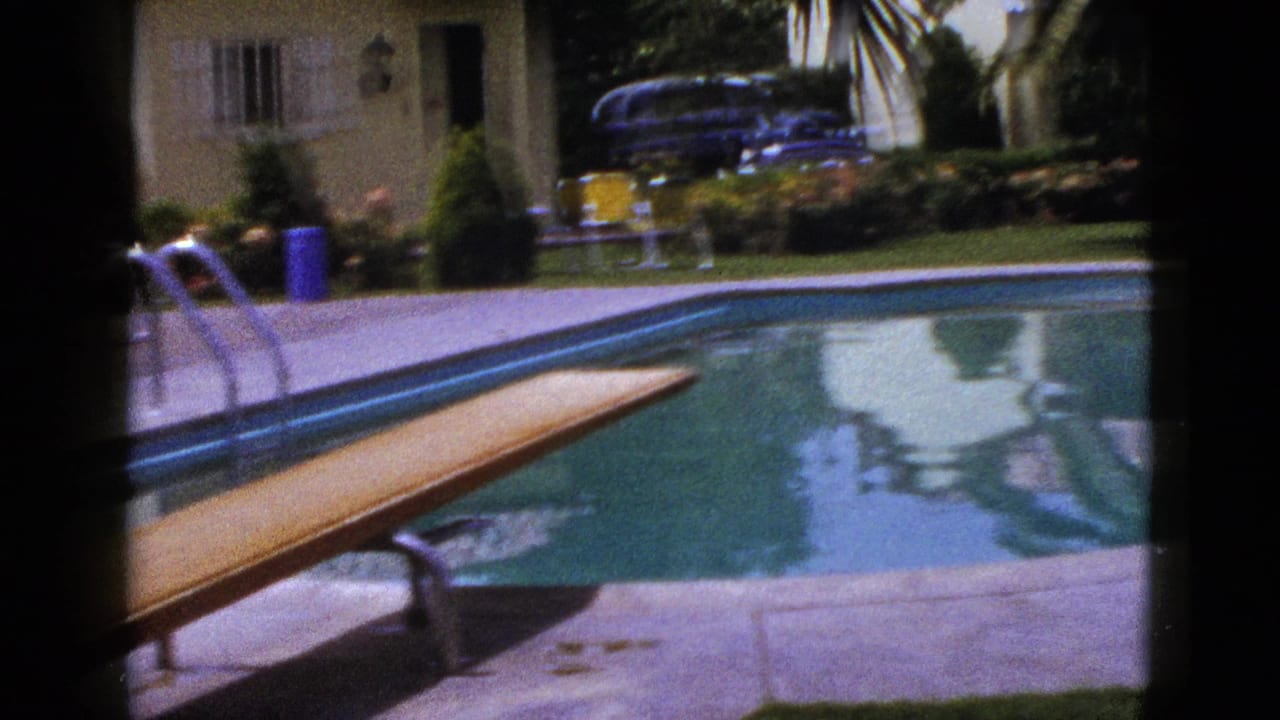 1953 New Mexico 8mm frame showing a backyard pool, wooden diving board, metal ladder, yellow house, lawn, planting, and dark classic car.