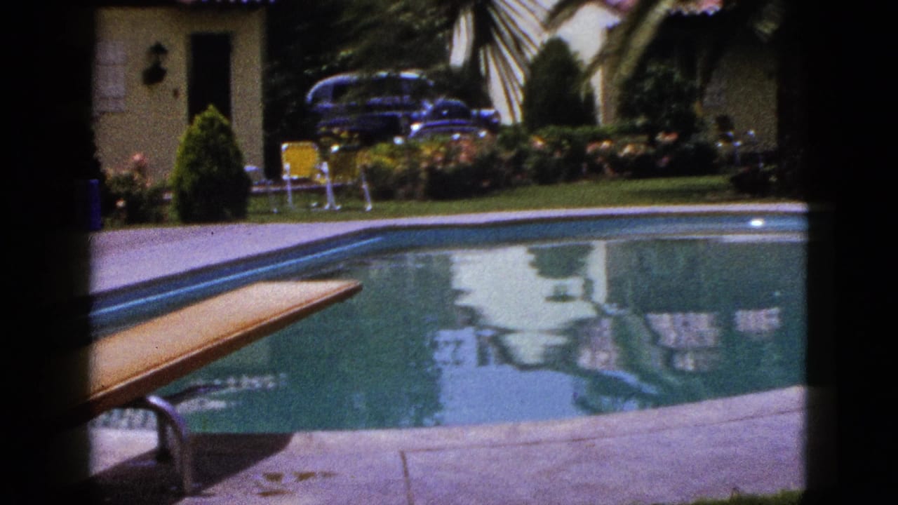 1953 New Mexico home-movie frame with the pool and diving board foregrounded, with lawn furniture, a house, planting, and classic car behind.
