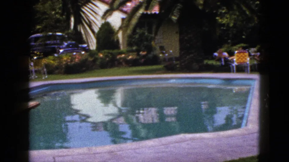 1953 New Mexico 8mm frame showing a wider poolside yard with a classic car, house, palm tree, lawn chairs, and distant seated people.