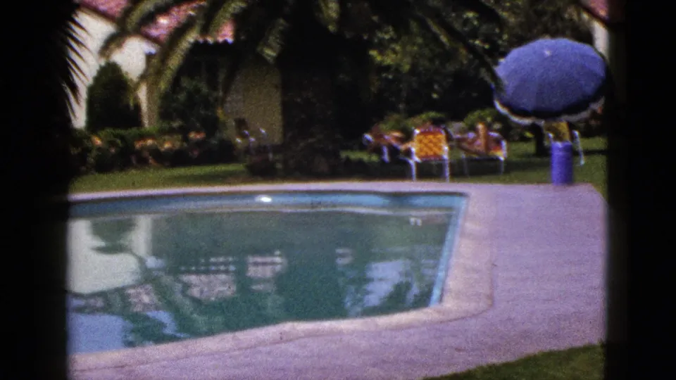 1953 New Mexico home-movie frame showing the pool, blue umbrella, lawn chairs, palm planting, house, and distant adults seated near the deck.