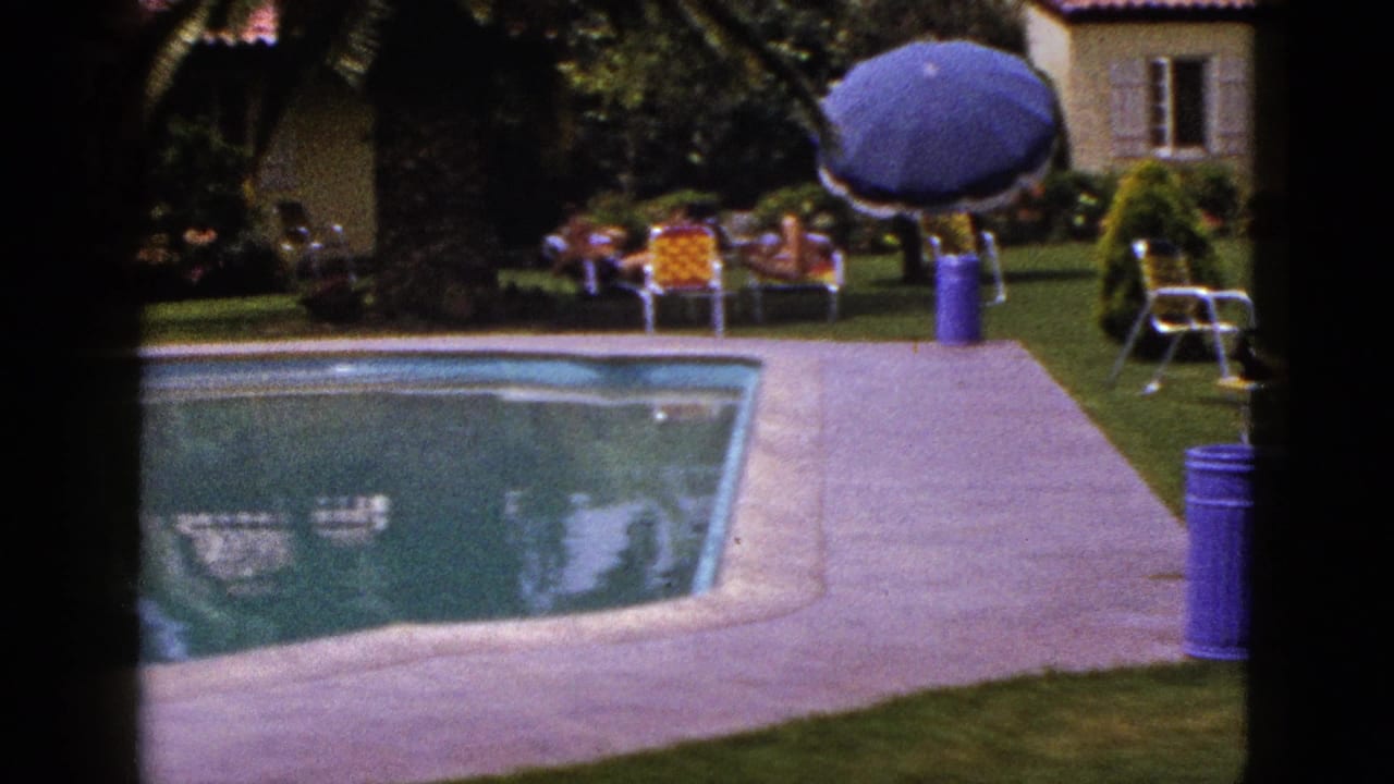 1953 New Mexico 8mm frame showing a backyard pool, concrete deck, lounge chairs, a blue umbrella and a stucco house with red tile roof.